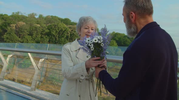 Elderly Man Holding Bouquet Behind Back and Gifting It to Senior Woman