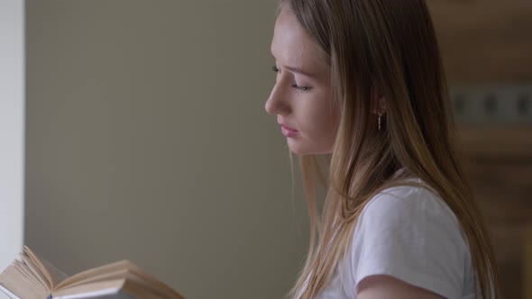 Side View of Young Absorbed Caucasian Woman Reading Book at Window Indoors