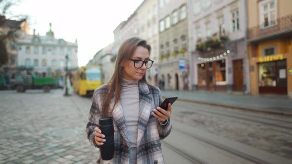 Woman in a Coat and Glasses with a Thermos Cup in Her Hands Talks on a Smartphone While Walking in