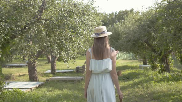 Attractive Young Woman in Straw Hat and Long White Dress Walking Through the Green Summer Garden