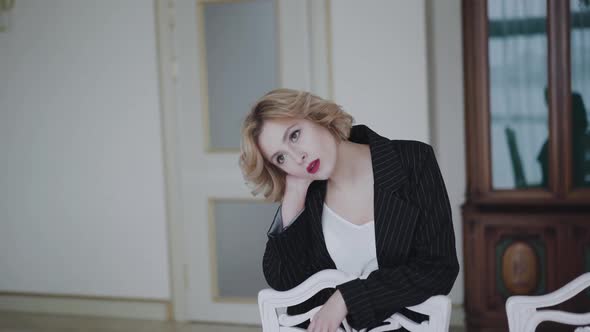 Portrait of Thoughtful Lady Leans on Chair and Poses at Camera in Luxury Home