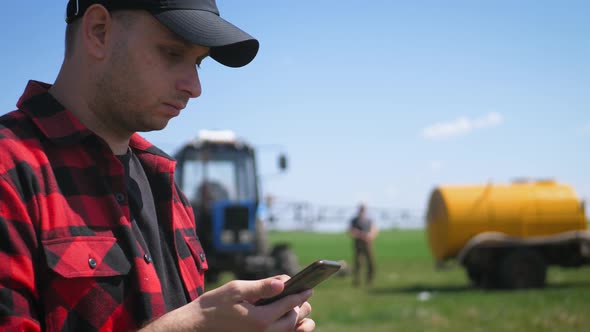 Young Attractive Farmer with Phone Standing in Field, Tractor Working in Green Field in Background