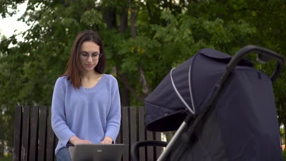 A Young Mother Works on a Laptop and Shakes Her Baby in a Stroller