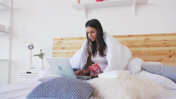 Сheerful Young Woman Using Laptop in Bed at Home Covered with Blanket and Eating Donut