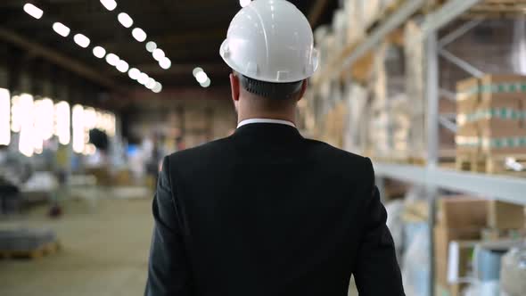 Back view of a boss in a suit and white helmet walking through a work warehouse