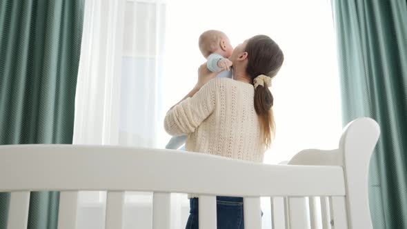 Young Mother Standing at Cradle and Holding Her Smiling Baby Son