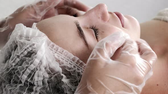 A female cosmetologist applies cream to the client's face with massaging movements.