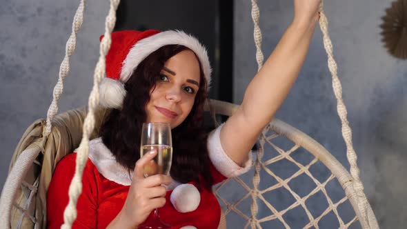 Young Woman in Santa Claus Suit with Glass of Champagne Sitting on Hammock