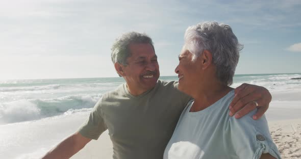 Happy hispanic senior couple embracing and walking on beach at sunset