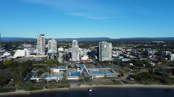 High panning view of a large Aquatic Centre on a coastal waterway with a towering urban city skyline