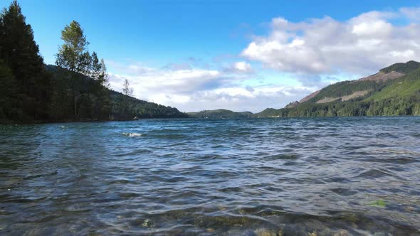 Clear water flying forward reveal lake distant rainforest green mountain hills with blue sky clouds