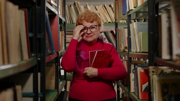 An Elderly Woman in Glasses Stands in the Library Between Bookshelves with Books in Her Hands