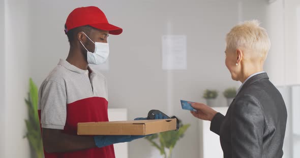 Deliveryman in Safety Mask and Gloves Delivering Pizza Box to Mature Businesswoman in Office