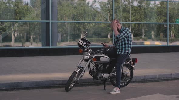 Young Man Motorcyclist with Smart Phone in Hands on Custom Motorcycle on Street