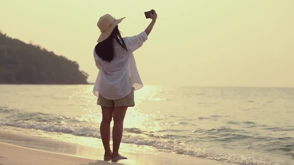 Asian beautiful young woman using smartphone taking a selfie while standing on the beach sunset.