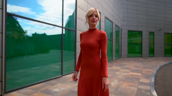 a Blonde Woman in a Red Dress and Black High-heeled Shoes Walks Along a Modern Building with Large