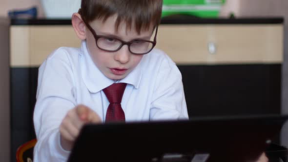 Caucasian schoolboy 7 years old in glasses at home doing homework holding a gadget in his hands