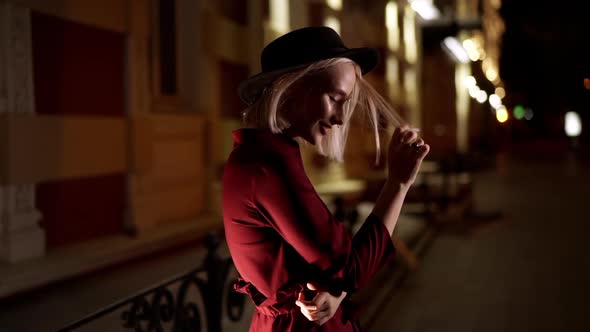 Portrait of Charming Woman with Blond Hairstyle and Hat Embarrassed and Flirting on Dark City Night