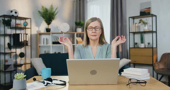 Senior Woman Meditating while Sitting at Table with Laptop