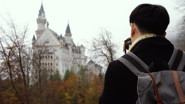 Male Asian Tourist with Backpack and Camera Standing and Enjoying View of Neuschwanstein Castle