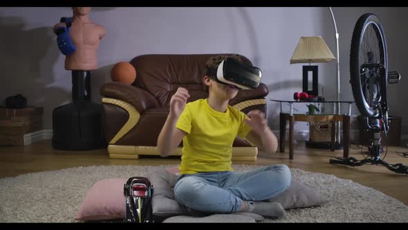 Portrair of Laughing Caucasian Boy in VR Googles Sitting on the Carpet and Moving His Body. Happy