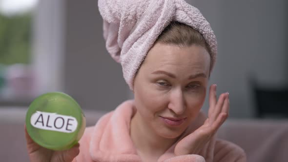 Confident Attractive Young Woman in Hair Towel and Bathrobe Applying Moisturizer on Face Looking at