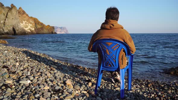 Back View of an Adult Male Anonymous Enjoying the Sea View While Sitting on a Blue Chair on a Spring