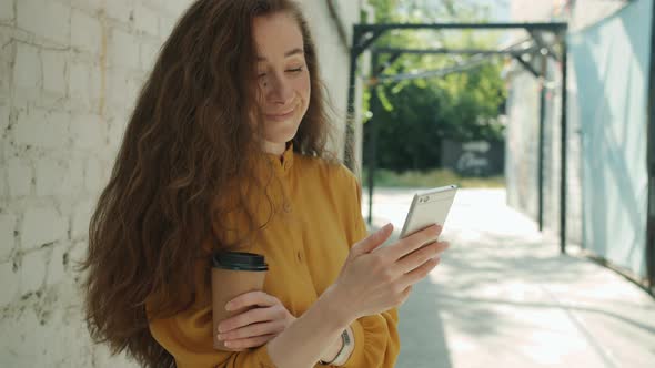 Goodlooking Young Woman Using Smartphone Smiling Holding to Go Coffee Standing Outside in City