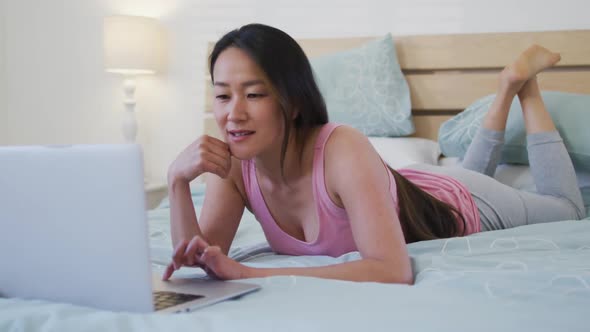 Happy asian woman lying on bed, using laptop in the morning