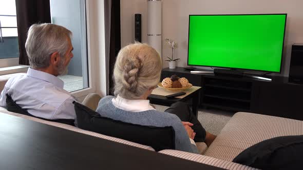 An Elderly Couple Sits on A Couch in A Living Room, Watches Tv with A Green Screen and Talks