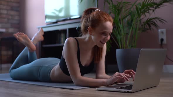 Portrait of Cheerful Young Woman Lying on the Floor and Using Laptop Computer at Apartment