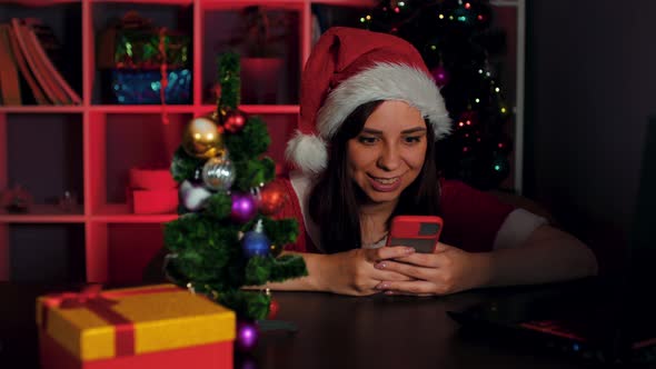 Young Woman in Santa Costume Using Smartphone in Office Sitting in Armchair in New Year's Atmosphere