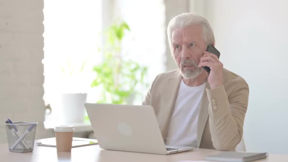 Old Man Talking on Phone While Using Laptop in Office