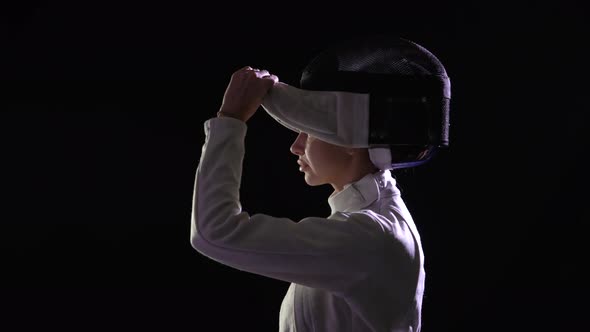 Side View of Young Female Fencer Putting Protective Helmet and Holding Fencing Rapier in Her Hands