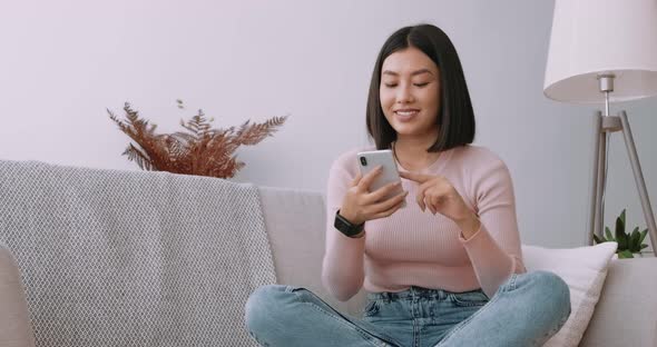 Young Asian Lady Surfing Social Media on Mobile Phone, Enjoying Amazing News on Sofa at Home