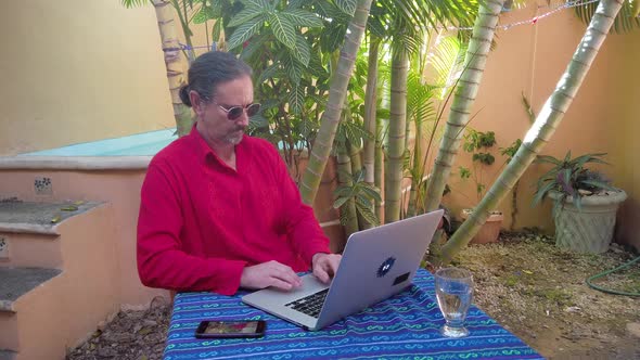 Mature man types on laptop keyboard at a table in a garden wearing a guayabera shirt.