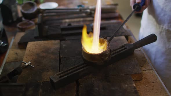 Caucasian female jeweller in workshop wearing apron, using gas burner, melting metal