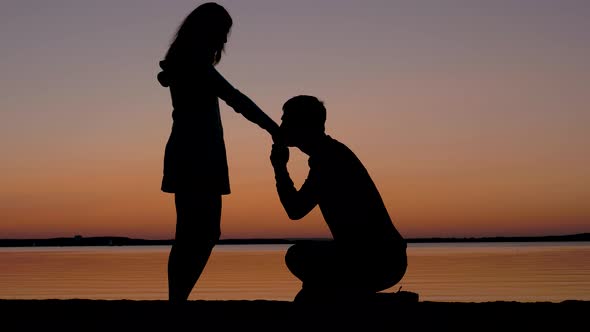 Man Sits On A Knee Then Takes The Woman Hand And Kisses Her At Sunset