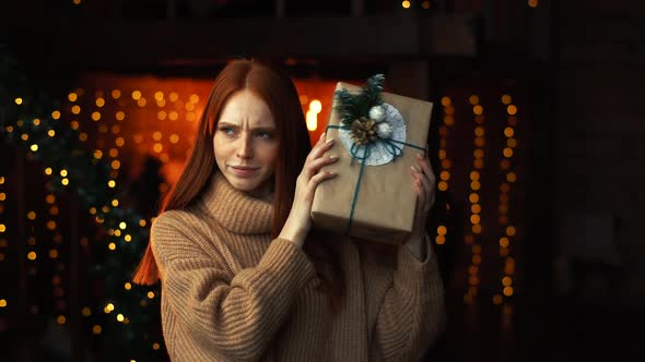 Portrait of Curious Beautiful Woman Shaking Gift Box with Christmas Presents Wrapped in Craft Paper.