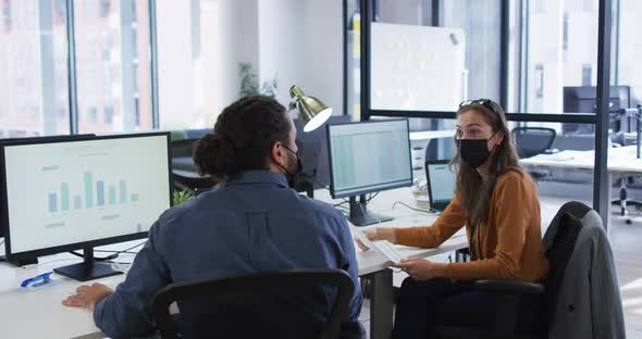 Diverse male and female colleague at desks using computers wearing face masks and elbow bumping