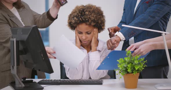 Exhausted Young African Businesswoman Sitting at Desk with Colleagues Shaking Papers in Her Face