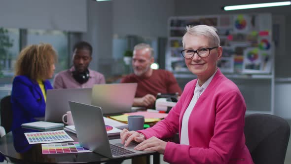 Successful Senior Businesswoman Smiling at Camera Sitting at Desk with Colleagues