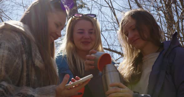 three young girls open a thermos and pour hot tea and laugh looking at the phone