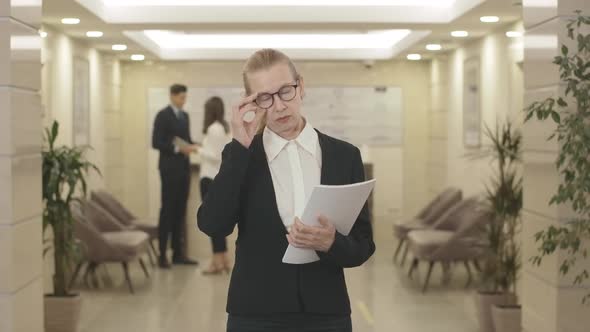 Portrait of Strict Mid-adult Businesswoman in Eyeglasses Posing with Documents in Hall. Confident