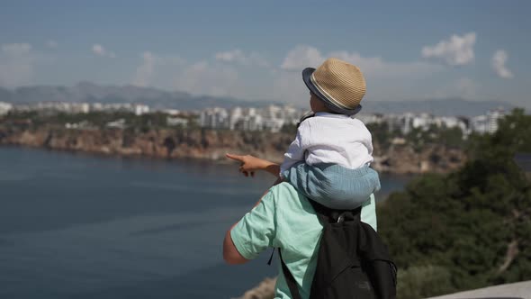 Mom Shows Toddler Sitting on Shoulders Views of Sea and Town