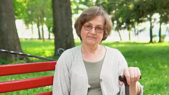 Portrait of Elderly Woman with Glasses and Cane Smiling on Bench in Public City Park