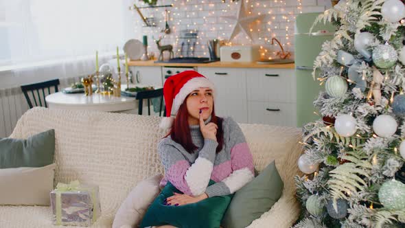 Young Woman in Santa Hat Sitting on Couch Near Christmas Tree in Living Room
