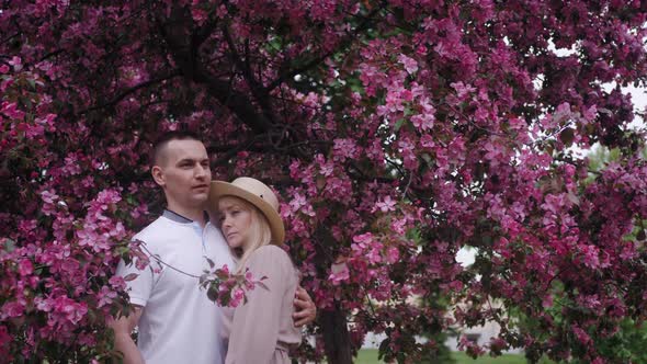 Romantic Beautiful Woman in a Hat Stands with a Man at a Cherry Blossom Tree in the Summer in the