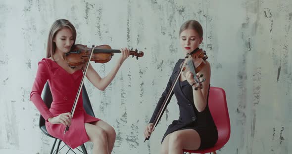 Two Elegant Girls in Dresses Playing the Violin Emotionally on Background Indoor