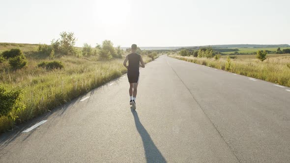 Rear View of Young Sportsman Jogging on a Road Out of City During Sunset